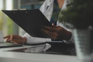 Serious female doctor using laptop and writing notes in medical journal sitting at desk. Young woman professional medic physician wearing white coat and stethoscope working on computer at workplace. photo