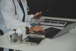 Serious female doctor using laptop and writing notes in medical journal sitting at desk. Young woman professional medic physician wearing white coat and stethoscope working on computer at workplace. photo