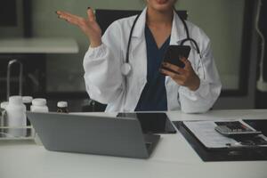 grave hembra médico utilizando ordenador portátil y escritura notas en médico diario sentado a escritorio. joven mujer profesional médico médico vistiendo blanco Saco y estetoscopio trabajando en computadora a lugar de trabajo. foto