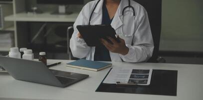 Serious female doctor using laptop and writing notes in medical journal sitting at desk. Young woman professional medic physician wearing white coat and stethoscope working on computer at workplace. photo