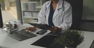 Serious female doctor using laptop and writing notes in medical journal sitting at desk. Young woman professional medic physician wearing white coat and stethoscope working on computer at workplace. photo