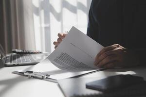 Asian businessman reviewing document reports at office workplace with computer laptop. legal expert, professional lawyer reading and checking financial documents or insurance contract photo