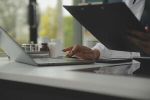 grave hembra médico utilizando ordenador portátil y escritura notas en médico diario sentado a escritorio. joven mujer profesional médico médico vistiendo blanco Saco y estetoscopio trabajando en computadora a lugar de trabajo. foto