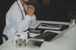 Serious female doctor using laptop and writing notes in medical journal sitting at desk. Young woman professional medic physician wearing white coat and stethoscope working on computer at workplace. photo