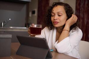 Attractive delightful woman reading news on digital tablet, browsing web sites, sitting at kitchen table over cup of tea photo