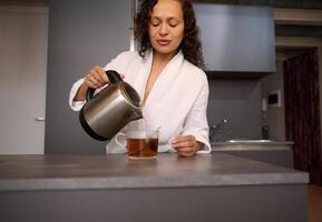 joven bonito mujer haciendo té para desayuno. detalles en hembra manos torrencial hirviendo agua desde un tetera dentro vaso taza con té bolsa. comida y bebida consumismo foto