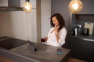 Multi ethnic woman in white bathrobe, making call from home interior, waving hello, greeting her interlocutor, smiling and holding a cup of hot tea in the morning, standing at kitchen counter photo