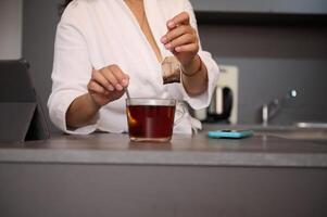 de cerca mujer en blanco bata de baño, edulcoración su té, en pie a cocina mesa con abierto digital tableta, en un minimalista hogar cocina interior foto