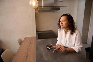 Attractive woman in white bathrobe, holding mobile phone while standing at table in the home, dreamily looking away photo