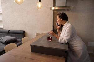 Relaxed young pretty woman in white bathrobe, talking on mobile phone, standing at kitchen table and mixing sugar in a cup of hot tea photo