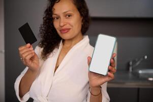 Young woman holds and shows at camera blank black credit card and smartphone with white empty mockup digital screen, makes online payments, transfers money via internet mobile banking or application photo