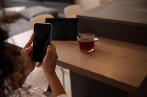 Close-up rear view of a woman sitting at table at home, texting on mobile phone with black mockup digital screen. Copy space for advertising text or mobile application photo