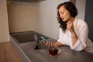 Beautiful young woman checking messages on digital tablet, sitting over a cup of hot herbal healing tea. photo