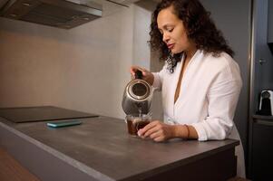 joven mujer haciendo té para desayuno. atractivo dama en blanco bata de baño torrencial hervido agua desde un eléctrico tetera dentro un vaso con bolsa de té foto