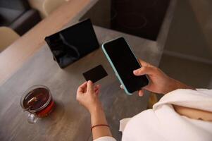 Close-up woman holds credit card, using smartphone with black mockup touch screen, online shopping. Businesswoman freelancer receives payment, checks balance on mobile app, booking internet tickets photo