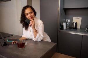 Middle aged confident beautiful woman holding bank credit card, using smartphone, online shopping, paying bills, receiving or making payment via mobile internet banking, standing at table at home photo