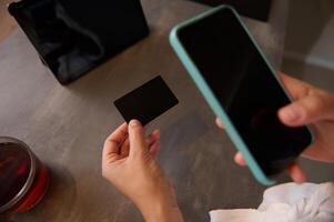 Close-up female hands hold a black mockup credit card with copy ad space for text, and using smart phone for online shopping, receiving or making payments via internet mobile banking application photo