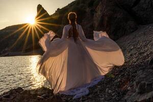 misterioso mujer silueta largo pelo camina en el playa Oceano agua, mar ninfa viento escucha a el ola. lanza arriba un largo blanco vestido, un adivinar puesta de sol. artístico foto desde el espalda sin un cara