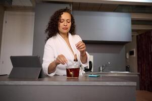 Confident young pretty brunette woman in white bathrobe, sweetening his tea, standing at kitchen table with open digital tablet, in a minimalist home kitchen interior photo