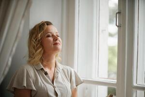 A middle-aged woman in a cream dress sits mysteriously and looks out the window on the windowsill. Green trees outside. photo