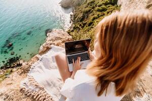 A woman is sitting on a rock overlooking the ocean with a laptop in front of her. She is enjoying the view and the peacefulness of the location. photo