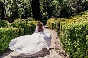 morena carreras blanco vestir parque. un hermosa mujer con largo marrón pelo y un largo blanco vestir carreras a lo largo el camino a lo largo el hermosa arbustos en el parque, posterior ver foto