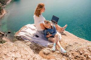 Lanza libre mujer con su hija trabajando en un ordenador portátil por el mar, mecanografía en el teclado, disfrutando el hermosa vista, destacando el idea de remoto trabajar. foto