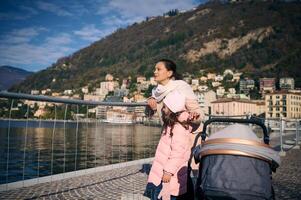 contento joven madre emprendedor paseante con pequeño niño, caminando con su pequeño niño niña en naturaleza, al aire libre, cerca lago de como en Italia foto