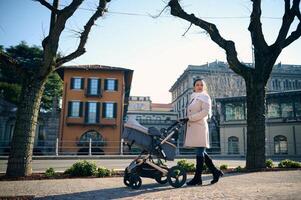 lleno longitud retrato de un joven elegante mamá, amoroso madre emprendedor un bebé paseante en el ciudad. bonito mujer caminando a lo largo el italiano ciudad como con su recién nacido bebé dormido en el bebé carro foto