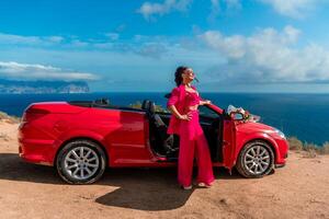 un mujer soportes en frente de un rojo convertible auto, participación un ramo de flores de flores concepto de libertad y aventura, como el mujer es vestido en un rojo atuendo y poses en frente de el coche. foto