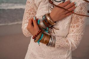 Model in boho style in a white long dress and silver jewelry on the beach. Her hair is braided, and there are many bracelets on her arms. photo