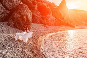 Mysterious woman silhouette long hair walks on the beach ocean water, sea nymph wind listens to the wave. Throws up a long white dress, a divine sunset. Artistic photo from the back without a face