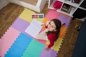 auténtico retrato de caucásico niño niña dibujo imagen utilizando vistoso pastel lápices de color, sonriente mirando a cámara, sentado en un rompecabezas alfombra a hogar. Arte y niños educación concepto. ver desde encima foto