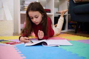Authentic portrait of Caucasian elementary age school child girl doing homework, drawing picture with colorful, pastel pencils, writing and reading at home photo