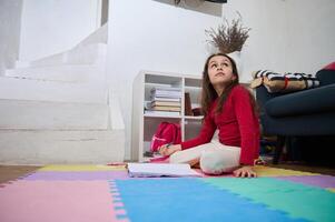 Adorable European little child girl in home clothes, sitting on a colorful puzzle carpet, dreamily looking aside, solving mathematics problems, writing essay while doing homework in her children room photo
