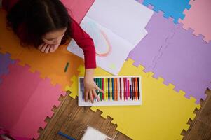 View from above of a little girl taking out colorful pencil from pencil case, drawing beautiful cloud with rainbow, lying on a multi colored puzzle carpet in cozy home interior. Kids education photo