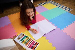 Top view of a cute little child girl lying down on a multi colored puzzle carpet at home, doing homework, drawing creative image with cloud and rainbow. Art and creativity. Kids education nd hobbies photo