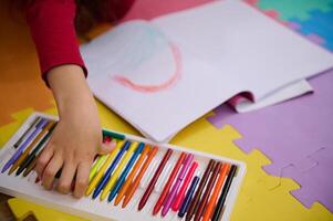 detalles en el manos de un pequeño niña tomando fuera vistoso lápiz desde lápiz caso, dibujo hermosa nube con arcoíris, acostado en un multi de colores rompecabezas alfombra en acogedor hogar interior foto