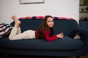 Caucasian cute elementary age school girl smiling looking at camera, lying on sofa, browsing websites, watching movies and having fun on her digital tablet with black mockup screen. photo