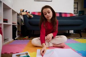Little child girl with long hair, sitting on the puzzle carpet at home, doing homework, studying, drawing picture with colorful pencils. People, kids education and entertainment. Art and creativity photo