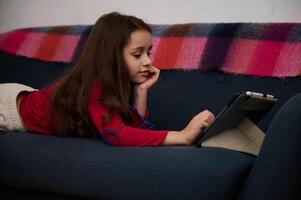 Adorable elementary age kid, cute little kid girl using digital tablet, watching movies while lying on the sofa at cozy home interior. Distance learning. Online education. Digital gadget addiction photo