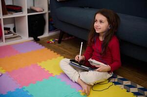 Smiling child girl doing homework on tablet, studying online sitting on multicolored puzzle carpet at home. Distance learning and kids education concept. Copy ad space on black mockup touch screen photo