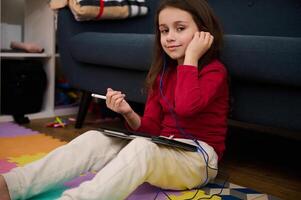 Portrait of a lovely child girl using digital tablet, studying online from home, smiling looking at camera, sitting on colorful puzzle carpet. Kids education. Distance learning. Autism. Diversity photo