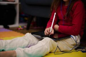 Close-up a white stylus of a digital tablet in hands of little child girl learning online, doing homework, drawing pictures, watching cartoons or listening to online webinar class. Kids and technology photo