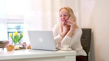 middle-aged adult woman working at computer talking on phone laughing drinking tea coffee light live communication morning breakfast flowers spring good joyful mood pleasant conversation easy life video