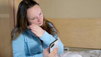ein Teenager Mädchen sitzt auf das Bett sieht aus beim das Telefon ein Weiß Fall mit grau Flecken ein jung Frau richtet sich auf ihr Haar will zu mögen zu nehmen ein Selfie Kommunikation online Internet video