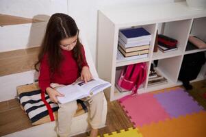 View from above school age child girl reading a book, sitting on steps of her room with multicolored puzzle carpet. The concept of back to school. Smart kids. Erudition and learning. World Book's Day photo