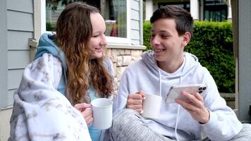 teen boy teenager girl sitting on porch drinking hot tea coffee looking at smartphone on shoulders plaid interest friendship watching movie Social networks discussing friends communication adolescence video