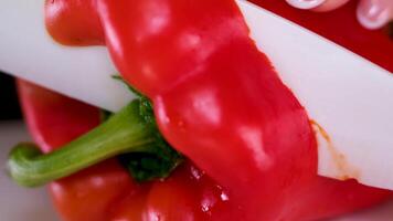 Professional cook in gloves cuts ripe red bell pepper with water drops on brown wooden board in restaurant extreme closeup video