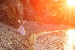 Mysterious woman silhouette long hair walks on the beach ocean water, sea nymph wind listens to the wave. Throws up a long white dress, a divine sunset. Artistic photo from the back without a face